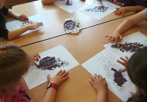 Grupa dzieci przy stoliku stempluje palcem umoczonym w fioletowej farbie gałązkę bzu.
